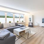 Wohnzimmer mit Sofa, Couchtisch und Fernseher. Große Fenster mit Blick auf Dach und Garten.
