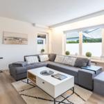 Wohnzimmer mit grauem Sofa, weißen Fenstern und Holzboden.