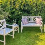 Weißer Gartenbereich mit Bank, Sessel und Tischen. Grüne Hecke im Hintergrund.