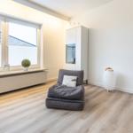 Ein Raum mit grauem Sitzkissen, weißen Wänden und Holzboden. Fenster mit Pflanzen und Schrank.