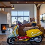 Ein gelbes Vespa-Motorrad steht im Wohnzimmer vor einem Kamin und großen Fenstern.