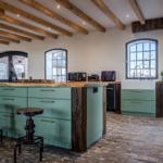 Küche mit grünen Schränken, Holzdecke und Steinboden. Barhocker und Fenster sind sichtbar.