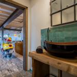 Bad mit Holzwaschtisch, grüner Fliesenwand und Spiegel. Durchgang zeigt Wohnbereich mit Motorrad.