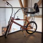 Ein orangefarbener Super Deluxe Fahrrad steht im Raum mit Holzboden und Balken.