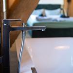 Duschbrause über weißem Badewannenrand, im Hintergrund Bett mit dunklem Kopfteil.