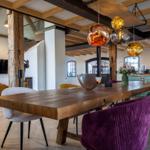 Essbereich mit massivem Holztisch und bunten Stühlen unter Holzbalkendecke.