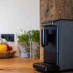 Eine schwarze Kaffeemaschine steht auf einer Holzoberfläche. Im Hintergrund sind Gemüse und Pflanzen zu sehen.