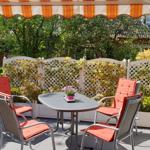 Terrasse mit Tisch, vier Stühlen und Sonnenschutz. Hintergrund: weißer Zaun und Pflanzen.