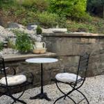 Garden with table and two chairs on gravel, stone wall and plants in background.