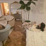 A person lies on a bed looking at their phone. In the background, there is a sofa and a mirror.