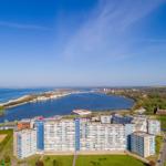 Panoramablick auf Wohnkomplex am Meer mit Hafen und grünen Flächen.