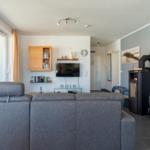 Wohnzimmer mit Sofa, Fernseher und Kaminofen. Große Fenster mit Blick auf Balkon.