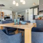 Essbereich mit Holztisch und blauen Stühlen. Küche und Wohnzimmer im Hintergrund.