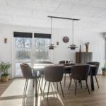 Dining area with table, chairs, and windows. Kitchen to the left. Wooden floor and white walls.