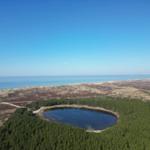Ein kreisförmiger See liegt inmitten von Wäldern, umgeben von Hügeln und dem Meer.