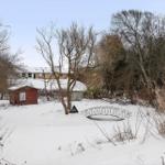 Snowy landscape with house, red shed, and metal bridge among trees.