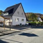 Haus mit dunklem Dach und weißer Fassade, gepflastertes Vorfeld und begrünte Umgebung.