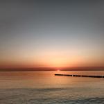 Sonnenuntergang über ruhigem Meer mit Holzpfählen im Vordergrund.