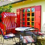 Roter Strandkorb und Sitzgruppe vor gelbem Haus mit roten Fenstern.