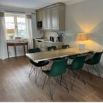 Essbereich mit langer Tisch und grünen Stühlen, Küchenzeile und Fenster mit Vorhängen.