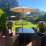 Terrasse mit Tisch, Stühlen und Sonnenschirm. Grünflächen und Blumen im Hintergrund.