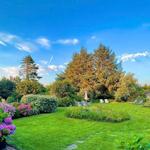 Grüner Garten mit Blumenbeeten, Liegestühlen und Bäumen unter blauem Himmel.