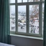 Durch das Fenster blickt man auf eine schneebedeckte Stadtlandschaft mit Dächern und Bäumen.