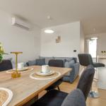 Dining area with table, chairs, and plant. Sofa and kitchen area in background.