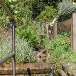 Ein Garten mit Brücke, Wasser und Pflanzen. Holzgeländer und Metallgeländer sind sichtbar.