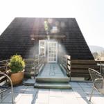 Haus mit Terrasse, Tisch, Stühlen und Pflanzen. Hintergrund: Berge und klarer Himmel.