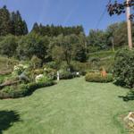 Großer Garten mit Rasenfläche, Blumenbeeten und Holzstuhl unter Bäumen.