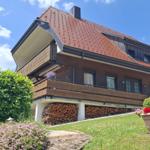 House with balcony, woodpile, and flowerbeds under blue sky.