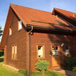 Holzhaus mit rotem Dach, Eingangstür und Fenstern, umgeben von Grünflächen und Bäumen.