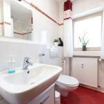 Badezimmer mit Waschbecken, Toilette und Dusche. Rote Fliesen und Fenster mit Vorhang.