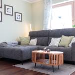 Ein Wohnzimmer mit grauem Sofa, Holztisch und Fenster mit Vorhängen.