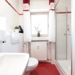 Badezimmer mit Dusche, Toilette und Waschbecken. Rote Fliesen und Fenster mit Vorhängen.