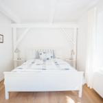 Ein Schlafzimmer mit weißem Bett, Holzboden und Fenster mit Vorhang.