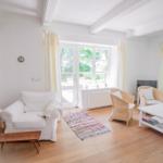 Wohnzimmer mit weißem Sofa, Rattanstühlen und Holzboden. Fenster mit Blick auf Garten.