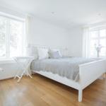 Schlafzimmer mit weißem Bett, Holzboden und zwei Fenstern mit weißen Vorhängen.