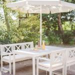 Weiße Terrassenmöbel mit Sonnenschirm auf gepflastertem Bereich unter Bäumen.