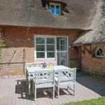 Terrasse mit weißem Tisch und Stühlen vor einem strohgedeckten Haus mit Backsteinmauer.