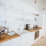 Küche mit Holzplatte, Spüle und weißen Schränken. Auf Regalen stehen Geschirr und Gläser.