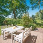 Terrasse mit weißem Tisch und Bänken, umgeben von Grün und Bäumen.