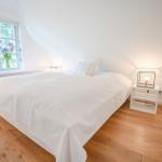 Schlafzimmer mit weißem Bett, Holzboden und Fenster mit Blumen.
