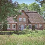 Rotes Backsteingebäude mit Strohdach und weißen Fenstern in grüner Umgebung.