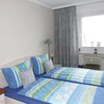 Zimmer mit Doppelbett, Fenster, Vorhängen und Nachttischen.