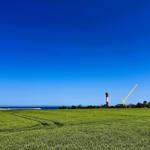 Rotes Leuchtturm mit weißem Streifen am Horizont über grüner Wiese und blauem Himmel.