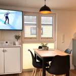 Essbereich mit Tisch, Stühlen und Fenster. TV zeigt Skifahrer. Küche daneben.