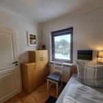 Schlafzimmer mit Bett, Schrank und Fenster. Wandlampe und Stuhl sind vorhanden.