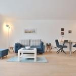 Wohnzimmer mit Sofa, Tisch und Stühlen. Holzboden und Fenster sichtbar.